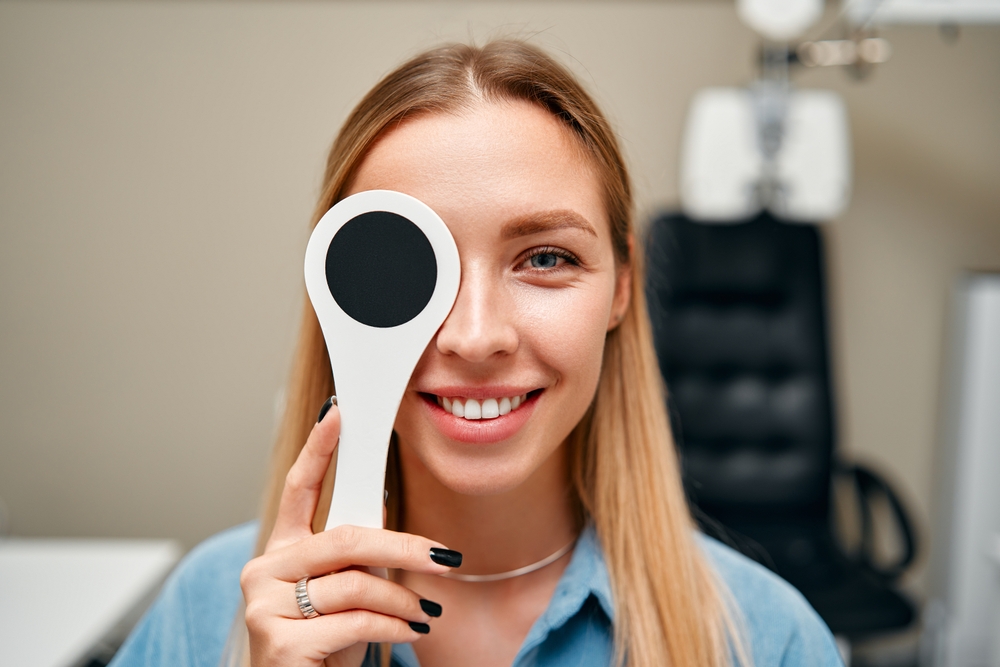 A smiling female patient holds an eye occluder during a professional vision test, demonstrating a thorough eye examination designed to assess clarity, focus, and overall ocular health - Comprehensive Eye Exams