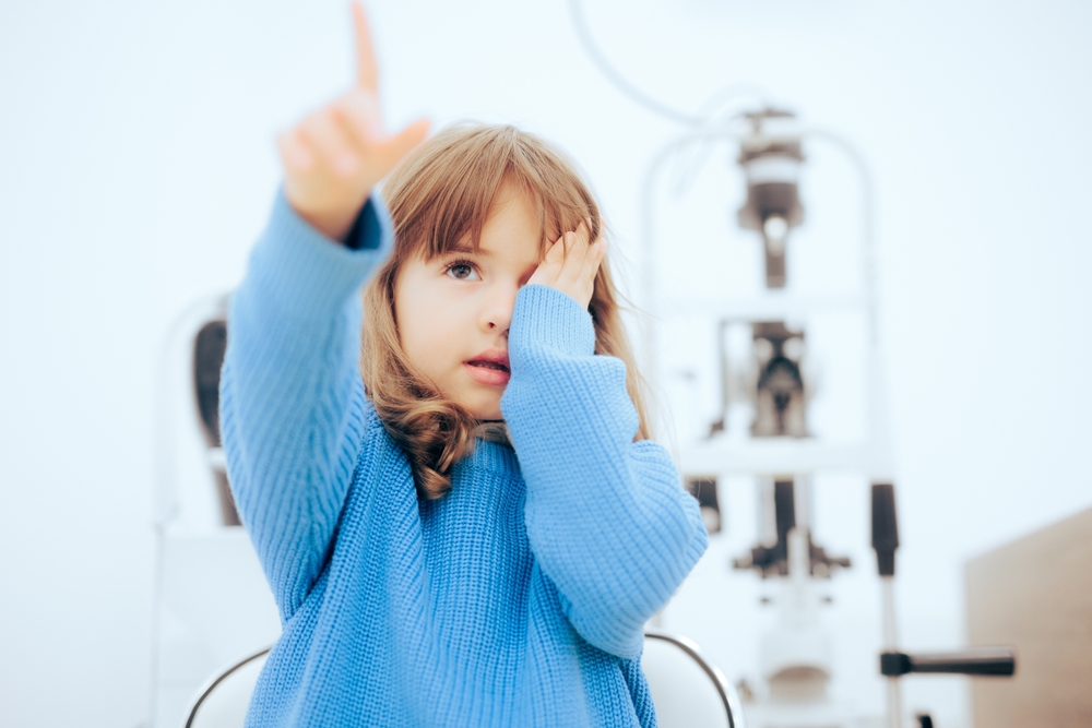 A young child participates in a pediatric eye exam by covering one eye and focusing during a vision screening, highlighting the importance of early detection and monitoring of visual development - Comprehensive Eye Exams