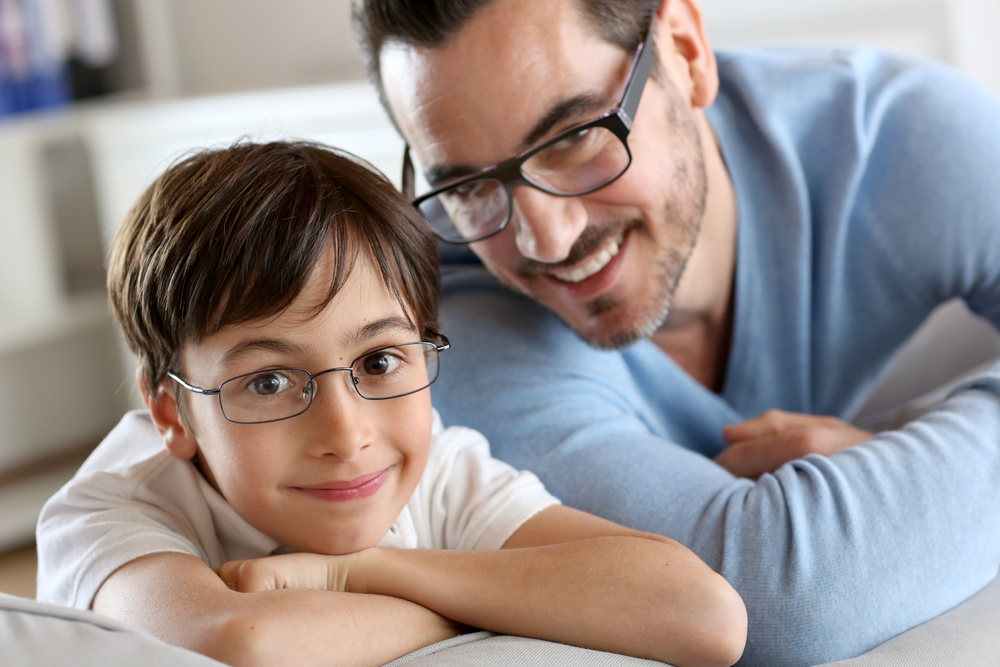 Father and son wearing eyeglasses and smiling indoors – Optometrist in San Francisco Happy father and son both wearing glasses and leaning on a couch, smiling warmly at the camera in a bright home setting – Optometrist in San Francisco